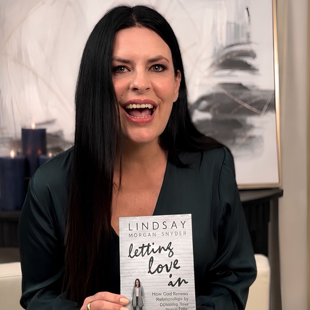 A woman with long dark hair, wearing a dark green top, smiles while holding a book titled Letting Love In by Lindsay Morgan Snyder. There are candles and abstract art in the background.