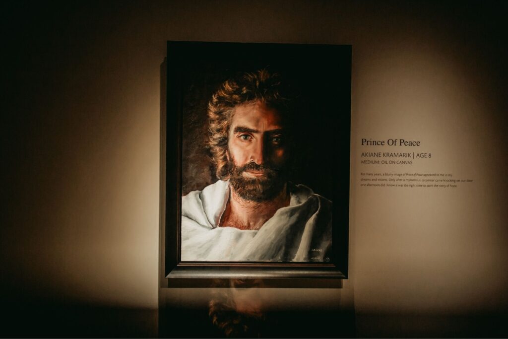 A framed painting of a man with long hair and a beard, draped in white, is displayed on a wall. Light focuses on the portrait. Text beside it reads Prince Of Peace and provides artist information.