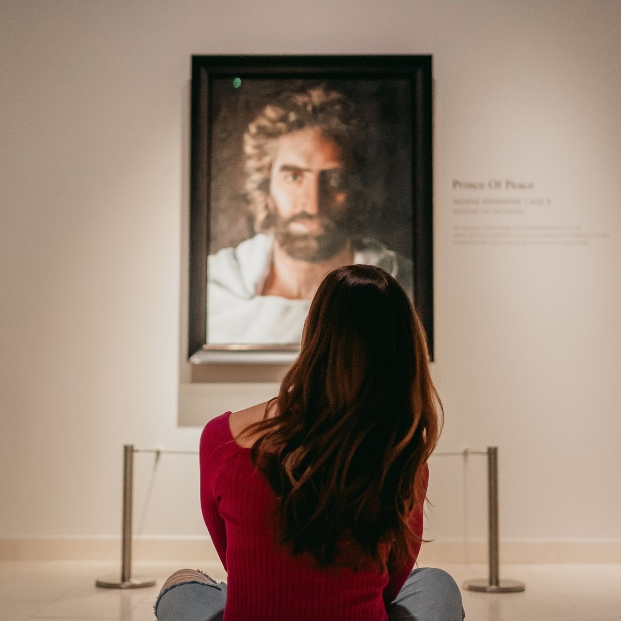 A woman with long brown hair in a red top sits on the floor, viewing a framed portrait of a bearded man displayed on a gallery wall.