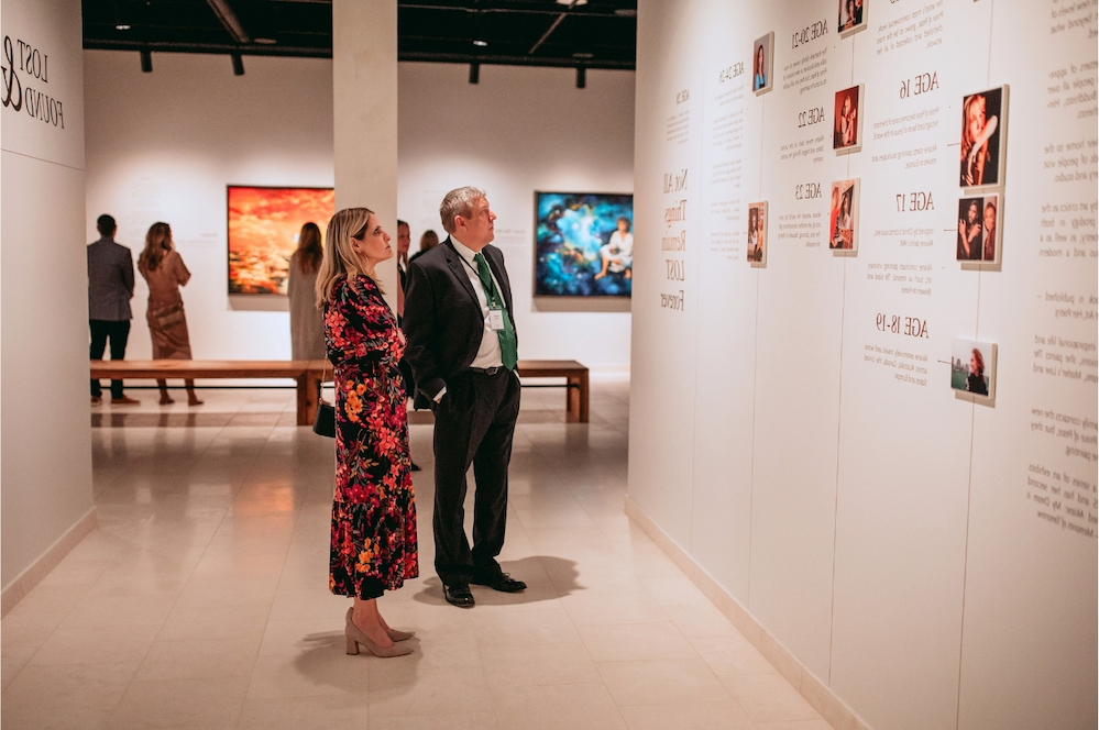 Two people stand in an art gallery, reading text on a white wall display. Other visitors and colorful paintings are visible in the background. The space is bright and modern.