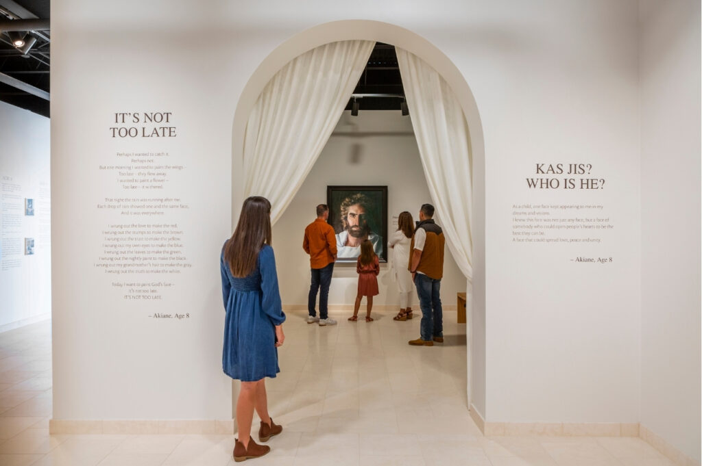 Five people stand in a gallery, looking at a portrait of Jesus through an arch with white curtains. Text is on the walls near the entrance.