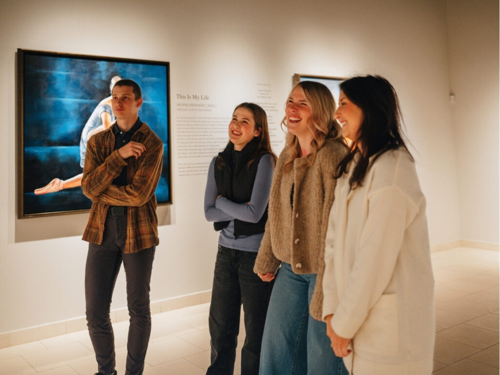 Four people stand smiling and talking in an art gallery, viewing a painting of a person in a seated pose on the wall behind them. The group appears relaxed and engaged in conversation.