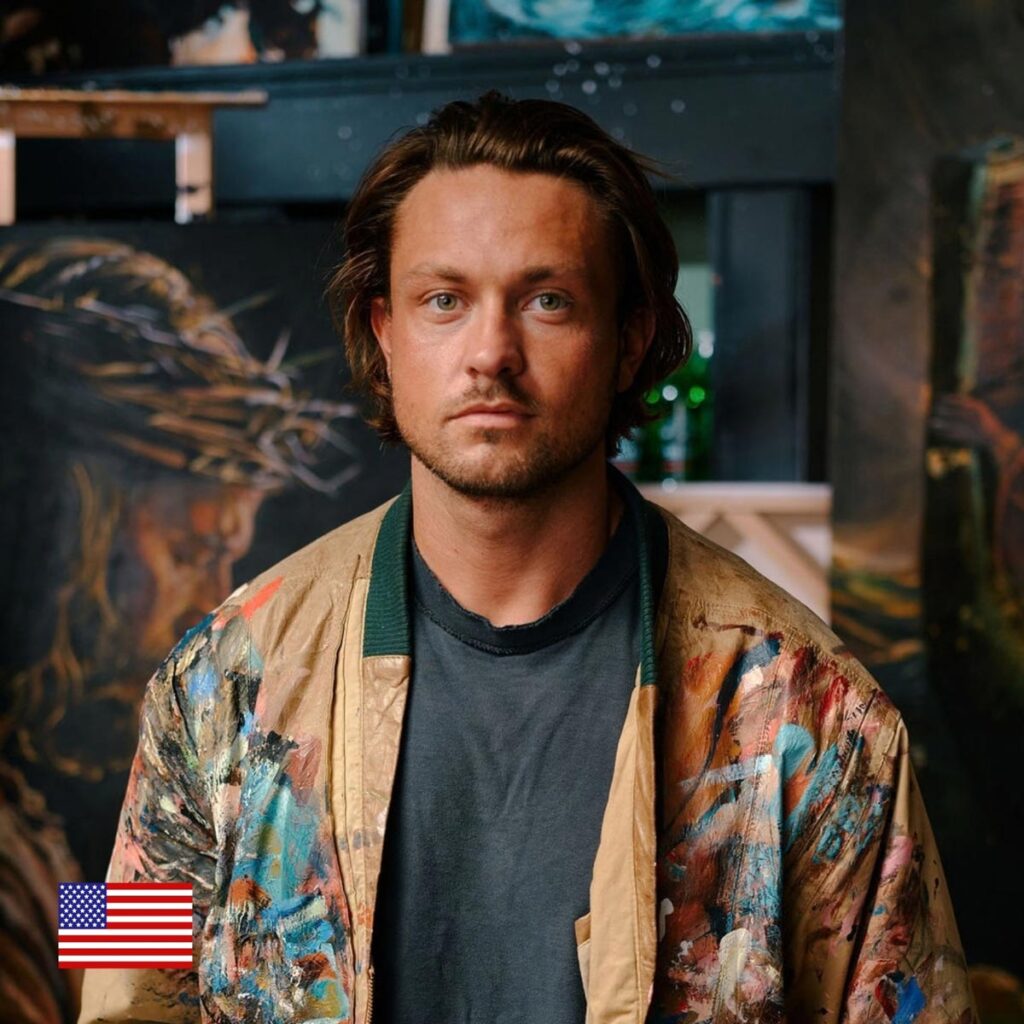 A man with shoulder-length brown hair and a serious expression wears a colorful, paint-splattered jacket. He stands in front of dark, artistic backgrounds. A small U.S. flag icon is visible in the lower left corner.