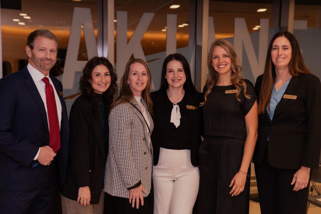 Six professionally dressed people—five women and one man—smile together indoors, standing in a row. The background shows large glass panels with partially visible text.