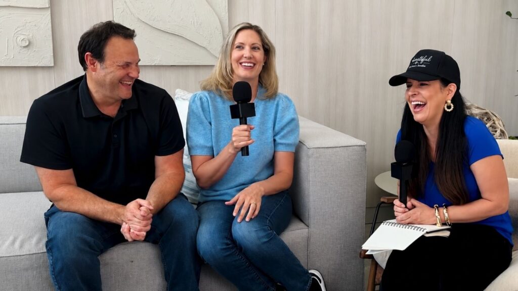 Three people sit on a couch, smiling and laughing during an interview. Two hold microphones, and one has a notepad and pen. The setting is casual and bright.