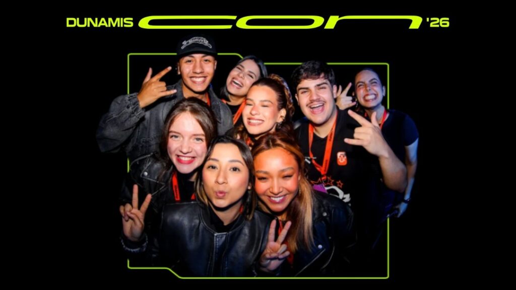A group of eight young adults smile and make peace signs for the camera at Dunamis Con 26, wearing event badges and dark clothing, with a black background and neon green event text above.