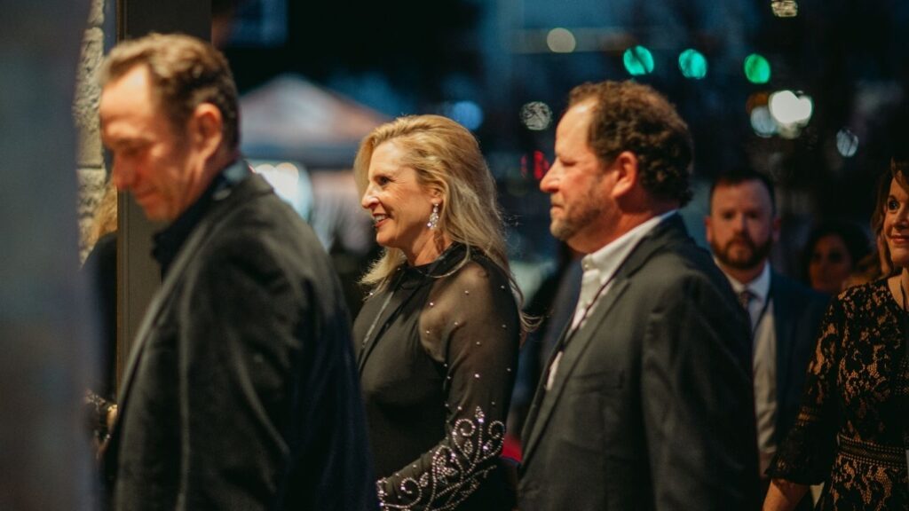 A group of well-dressed people, including a smiling woman in a black dress, stand in line at an evening event. The background is softly lit and slightly blurred.