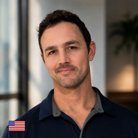 A man with short dark hair and light stubble smiles softly, wearing a navy collared shirt. An American flag icon appears in the lower left corner. The background is softly lit and slightly blurred.