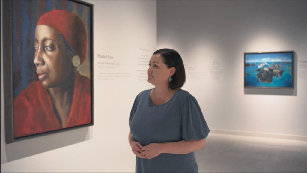 A woman in a blue top stands in an art gallery, looking thoughtfully at a portrait of a woman in a red headscarf. Another painting of a blue seascape hangs on the wall in the background.