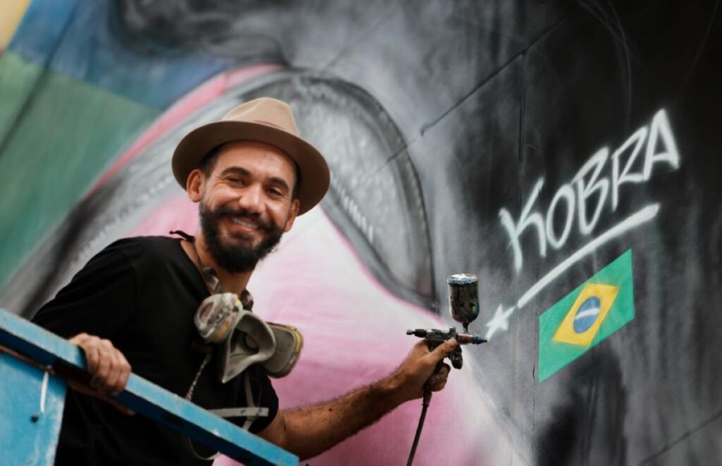 A smiling artist with a beard and hat, holding a spray can, stands beside a mural with the word KOBRA and a Brazilian flag painted on it.
