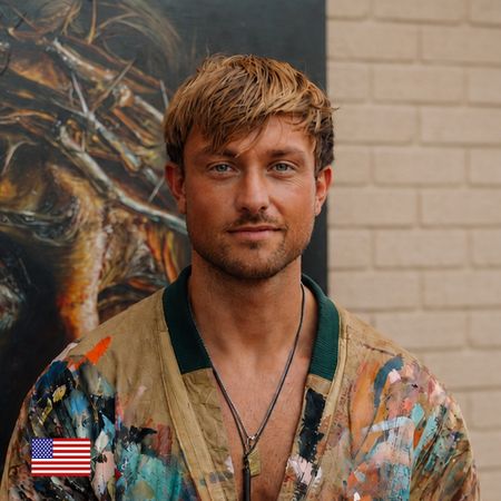 A man with blond hair in a colorful, paint-splattered jacket stands in front of a brick wall and an abstract painting, with a small U.S. flag icon in the bottom left corner.