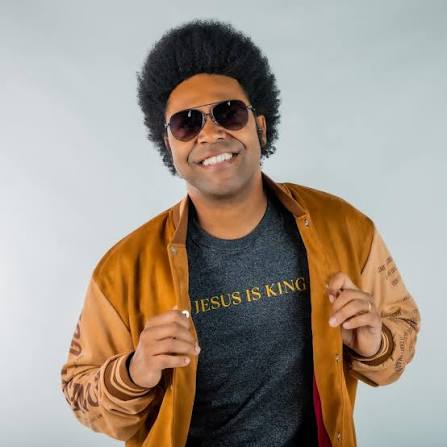 Smiling man in sunglasses with an afro hairstyle, wearing a brown jacket over a shirt that says JESUS IS KING, standing against a light grey background.