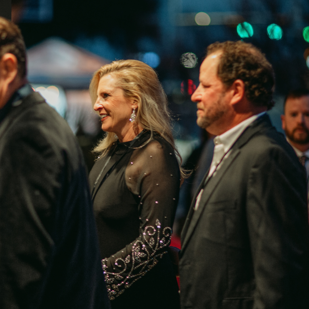 A woman in a black dress with jeweled sleeves and a man in a suit walk together at an evening event, with blurred people and lights in the background.