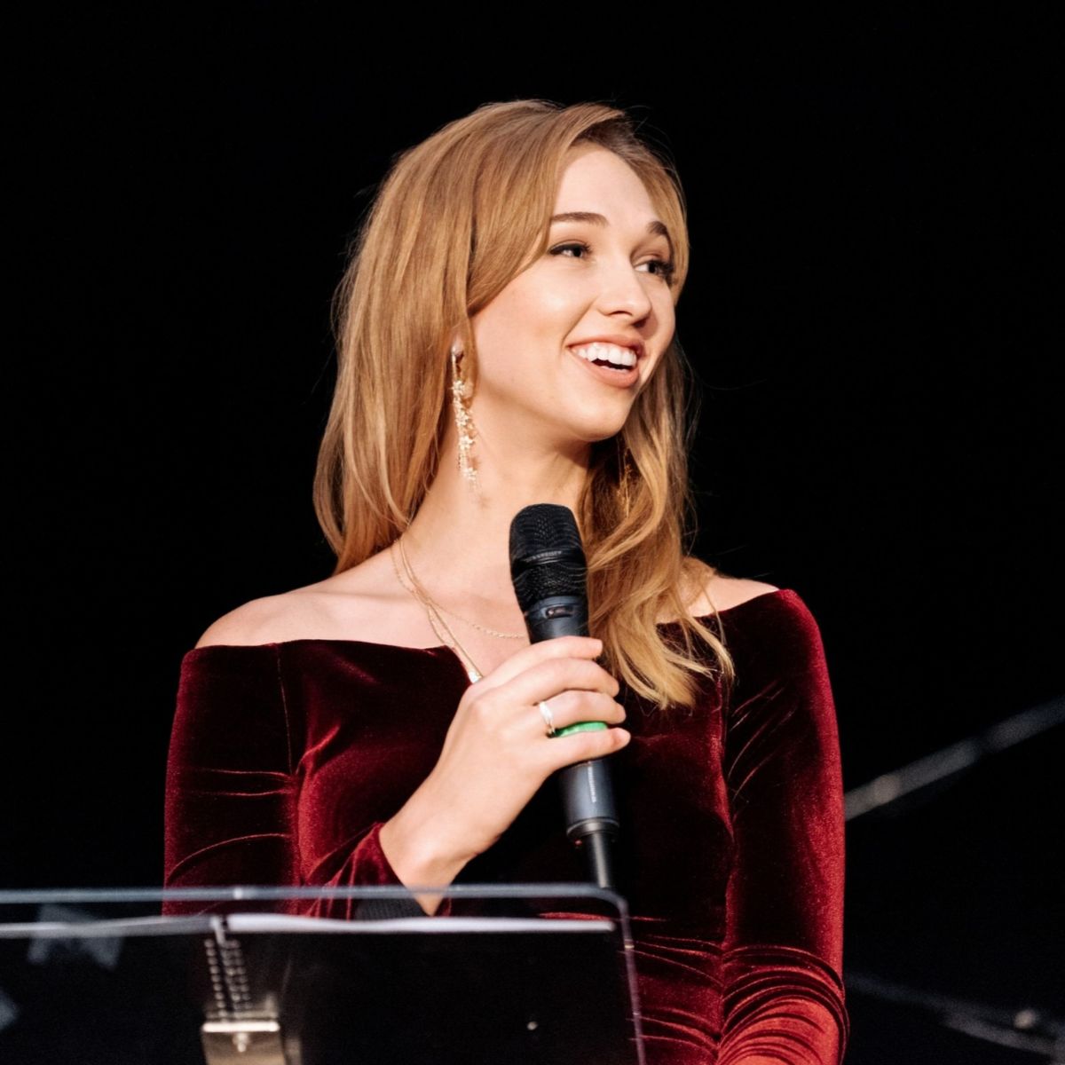 A smiling woman in a dark red dress stands at a podium holding a microphone.