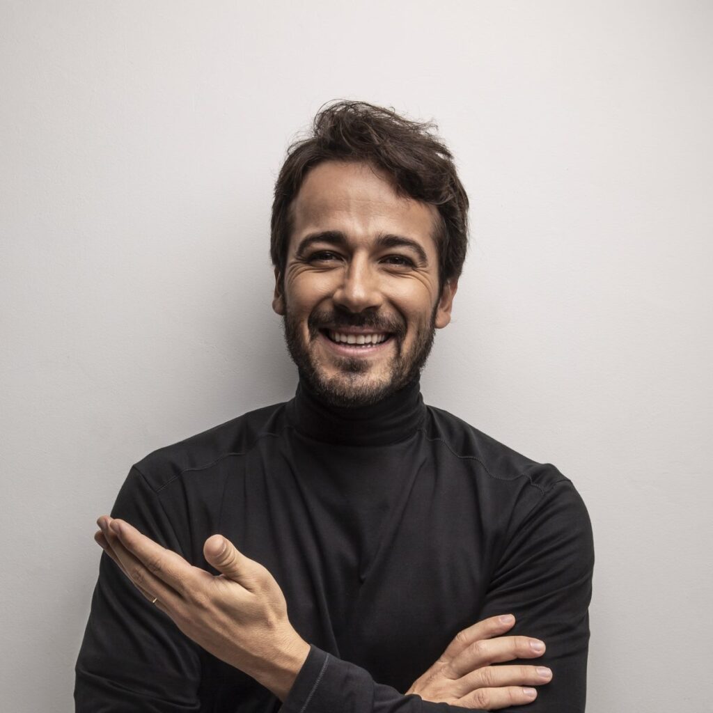 A smiling man with a beard in a black turtleneck stands against a light gray wall, arms crossed with one hand open.